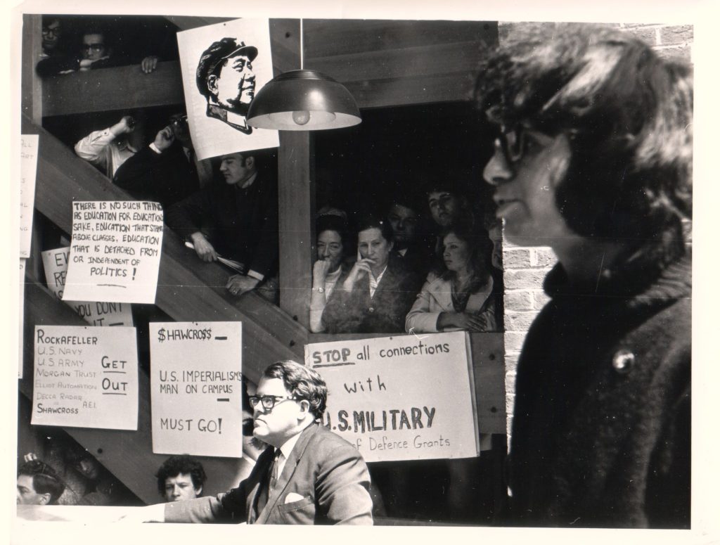 A sepia toned black and white photograph of Professor Asa Briggs sitting in Falmer House Common Room surrounded by student protesters and anti-imperialist posters