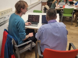 A photograph showing two people working together at laptops.  The facilitator (George) is demonstrating on one device and watching the colleague complete the same activity on theirs.