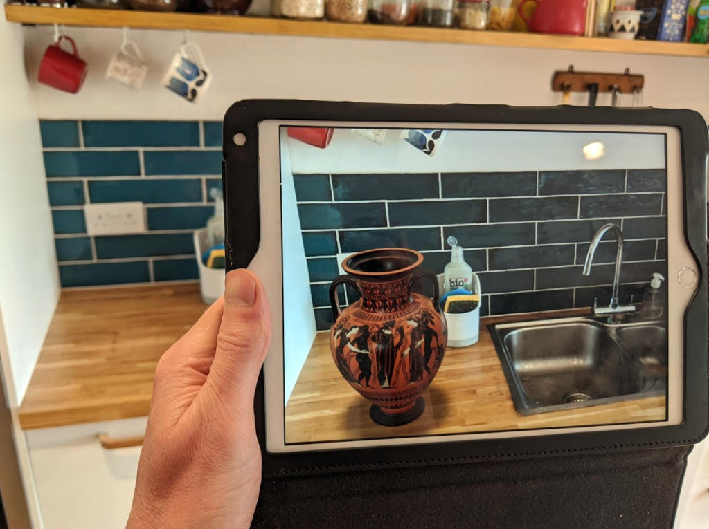 Image shows a hand holding an iPad. on the screen of the iPad a 3D model of an amphora is overlaid on the scene captured by the iPad camera. 