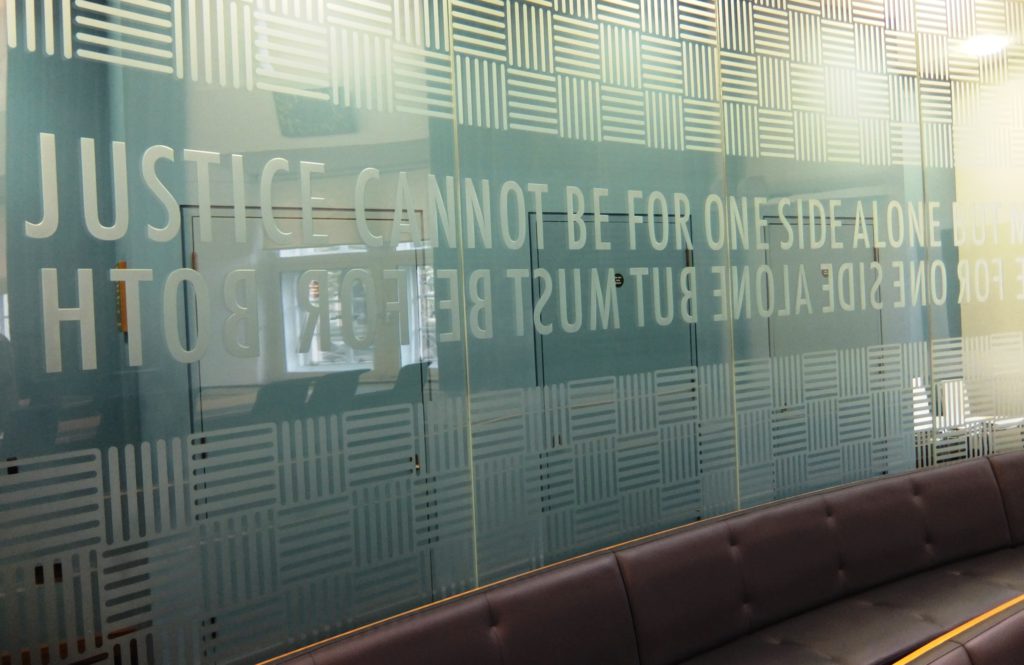 picture of Court 2 - the glass wall allows visitors to stand behind it or sit in the public benches in front. The screen is etched with the words: “Justice cannot be for one side alone but must be for both”. 