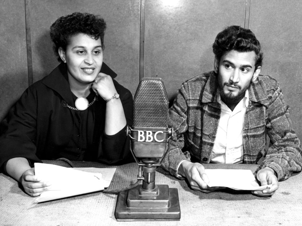 Pauline Henriques is seated at a desk with Samuel Sevlon in this black and white image. A large microphone bearing the letters BBC is inbetween them. They both hold pages of paper in front of them.  