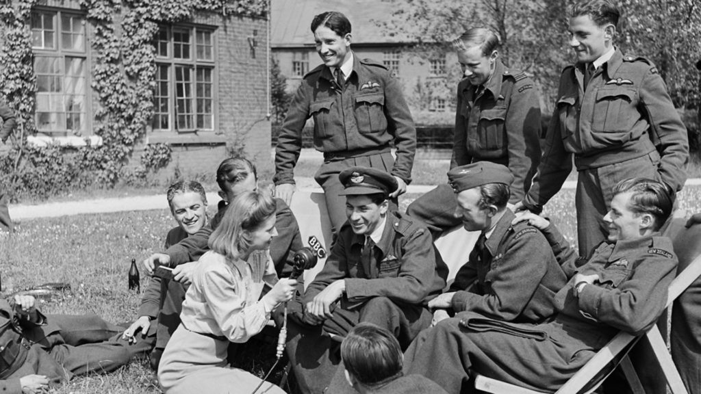 BBC reporter Noni Wright is seated on the grass, holding a microphone. A young man is speaking to her while 8 others look on. 