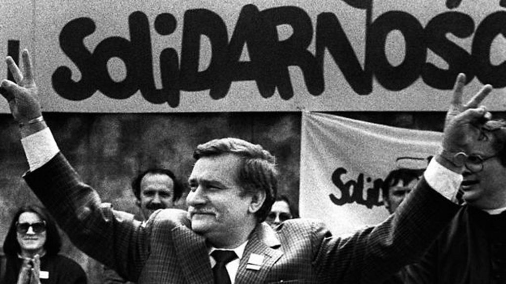 A man is standing with his arms raised triumphantly. Behind him is a large banner bearing the word Solidarity in Polish. 