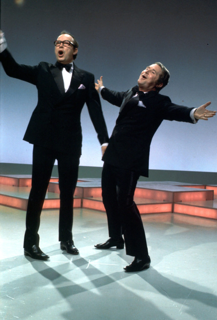 Morecambe and Wise are standing on a stage, performing. Morecambe is singing, one arm raised, while Ernie has both arms outstretched, and is leaning back. Both are wearing black suits and bow ties.