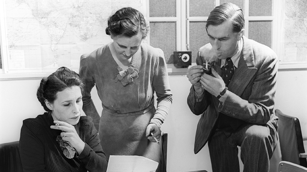 A woman is seated at a desk holding a sheet of paper. A woman is standing behind her, reading the document, alongside a man, who is lighting a pipe. The women are both holding cigarettes