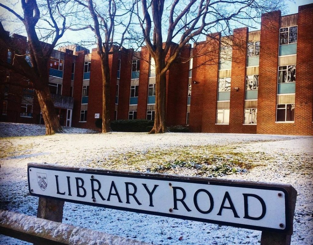 The Institute of Development Studies building (1960s redbrick design) in winter with a light dusting of snow on the lawn and a 'Library Road' sign in the foreground, at the University of Sussex campus in Falmer.