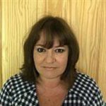 A woman (Jo Tregenza) with shoulder-length brown hair wearing a black-and-white checkered shirt, standing in front of a light wooden panel background