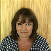 A woman (Jo Tregenza) with shoulder-length brown hair wearing a black-and-white checkered shirt, standing in front of a light wooden panel background