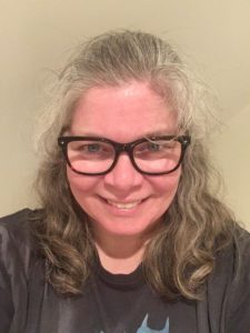 A person wearing glasses, and with long, wavy, grey-and-brown hair is shown from the shoulders up, wearing a dark T shirt, against a plain background.