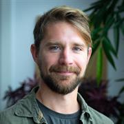 A photograph of a bearded man with fair hair, wearing a green shirt and t-shirt