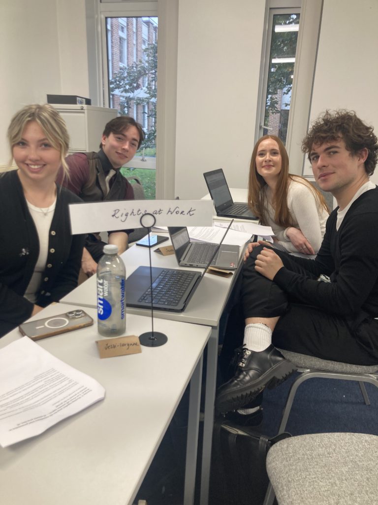 students of diverse origins sitting around tables in a seminar room, dressed in smart casual clothes. They have laptops in front of them and the name of their law firm at the front of the photo. They are smiling at the camera.