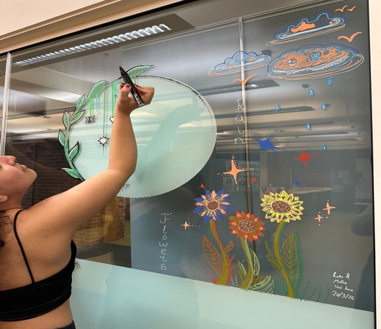A student draws a window painting on the glass windows looking into the Wellbeing area.