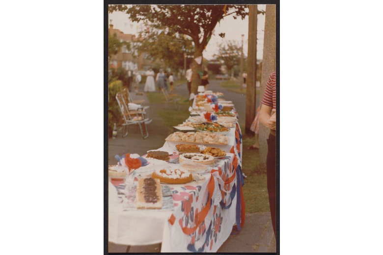 A scan of a colour photograph of a long picnic table. It is covered in a white table cloth and red, white and blue decorations. It is covered in food, forming part of a street party.