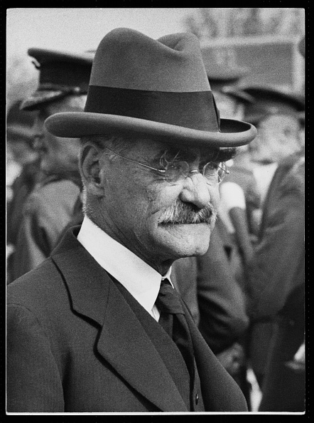 A side portrait of Rudyard Kipling in a crowd of officers wearing a suit and tie and a hat