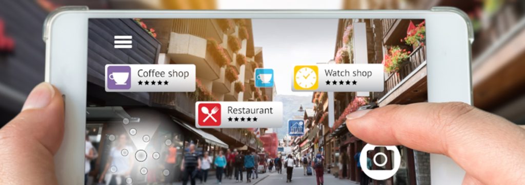 A mobile phone is held in two hands. On  the screen is an augmented view of a street with pop up icons of different venues, 'coffee shop', 'restaurant', 'watch shop'.