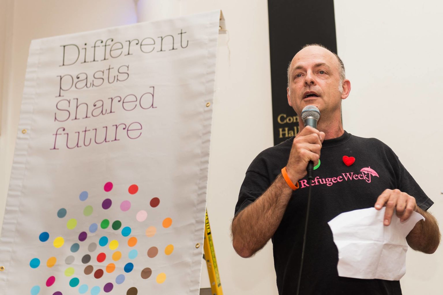 Richard Williams, a white man in a black tee-shirt bearing the legend 'Refugee Week' stands talking into a microphone next to a banner reading 'Different pasts, Shared future'.