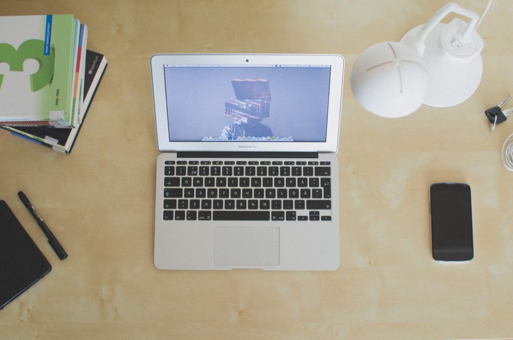 An aerial view of a desk with a laptop in the centre surrounded by a stack of books, a lamp, keys, a phone, a pen and a notebook.