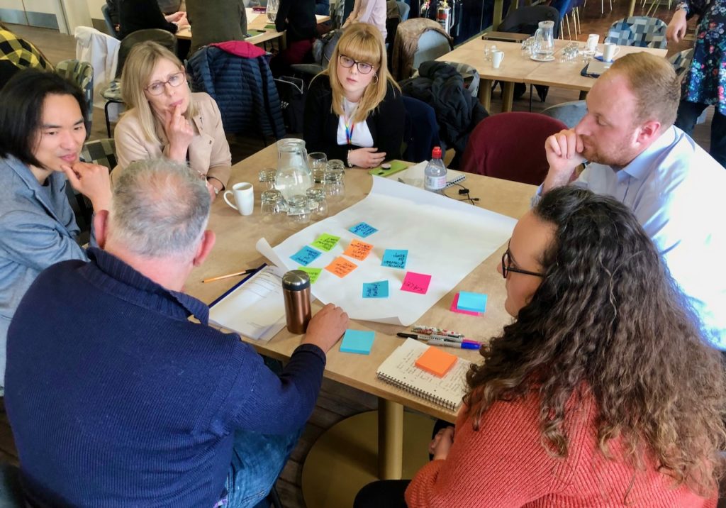 Group of 6 event attendees sitting around a table in discussion with post-it notes.