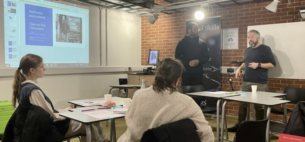 Four people in a classroom, with a projection screen on the left. The two standing people are presenting their work to the two colleagues sitting down.