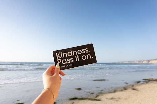 Hand outstretched from camera pod, holding a small black card with white text, which reads: Kindness, pass it on. Background is sandy beach, the sea and a blue sky.