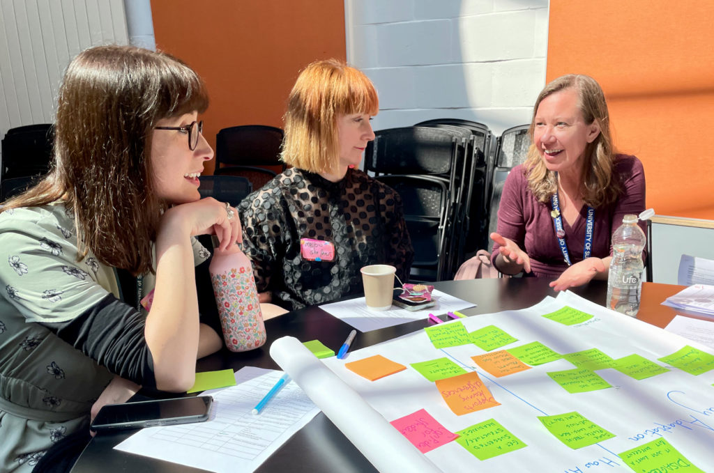 A photo of three participants in discussion at the 2023 Sussex Education Festival. They are sat at a table with many post-it notes on it, presumably generating ideas together.
