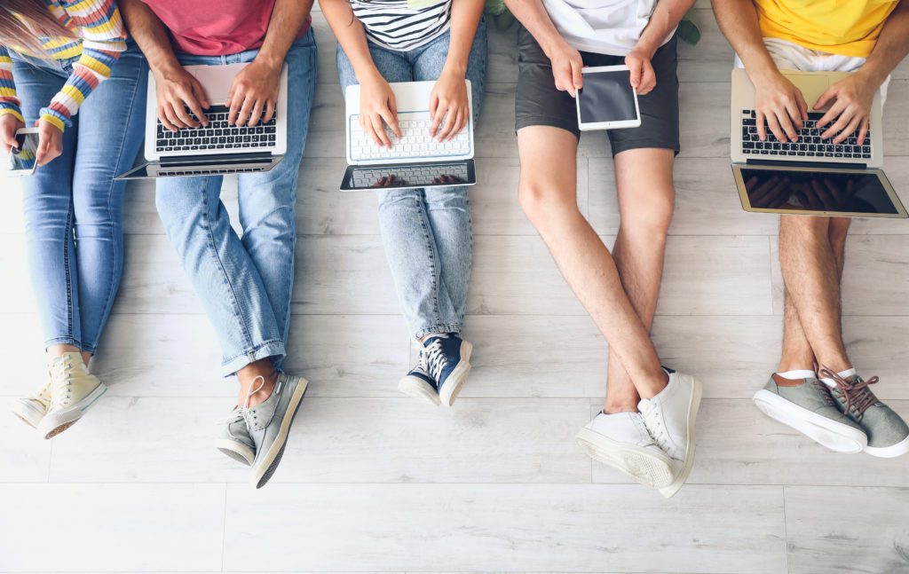 A photo of five university students, sitting using their devices. The photo is from above, framing the devices and the students' legs as they sit side-by-side on the floor. This highlights their diverse clothes, backgrounds and the devices they use to get online.