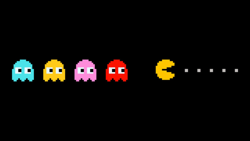 An image from the game Pacman. The yellow circle character of pacman is eating white dots followed by colourful ghosts. 